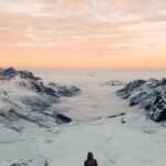 Elevation Gain - Person Standing in Snowy Field Under White and Orange Skies